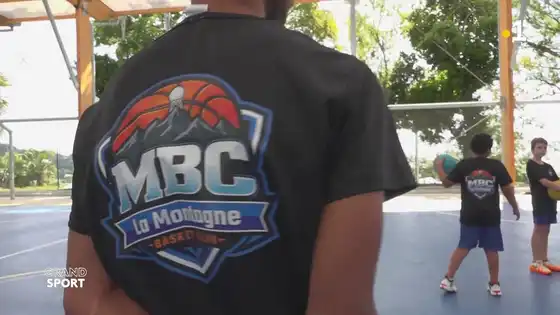 Image du reportage de Réunion la 1ère : un joueur de dos, en t-shirt noir au logo du MBC La Montagne Basket Club, devant des enfants à l'entraînement sous le plateau couvert de Ruisseau Blanc.