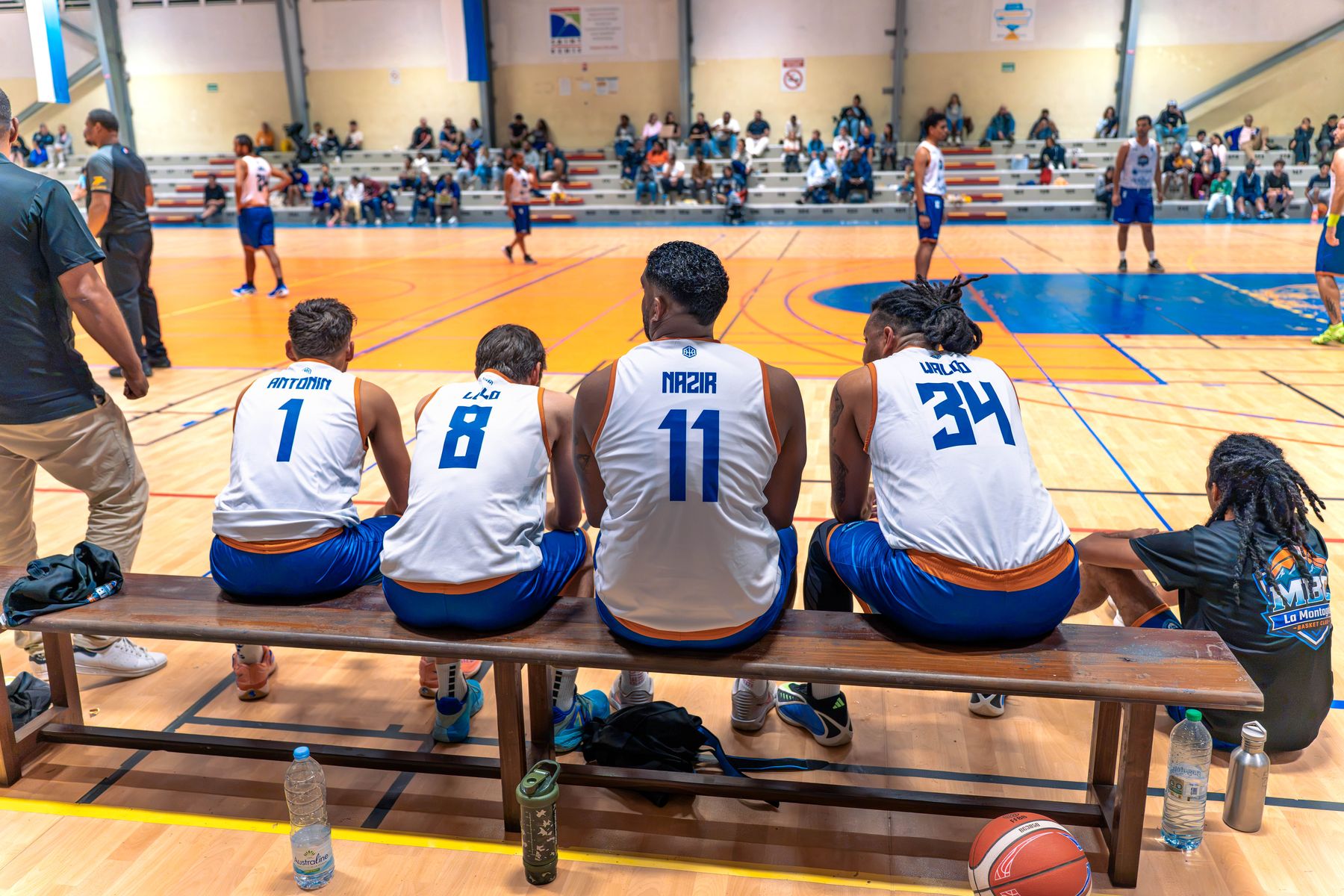 Le banc du MBC vu de dos : les numéros 1, 8, 11 (Kevin Nazir) et 34, assis face au terrain et aux tribunes du gymnase de La Montagne.
