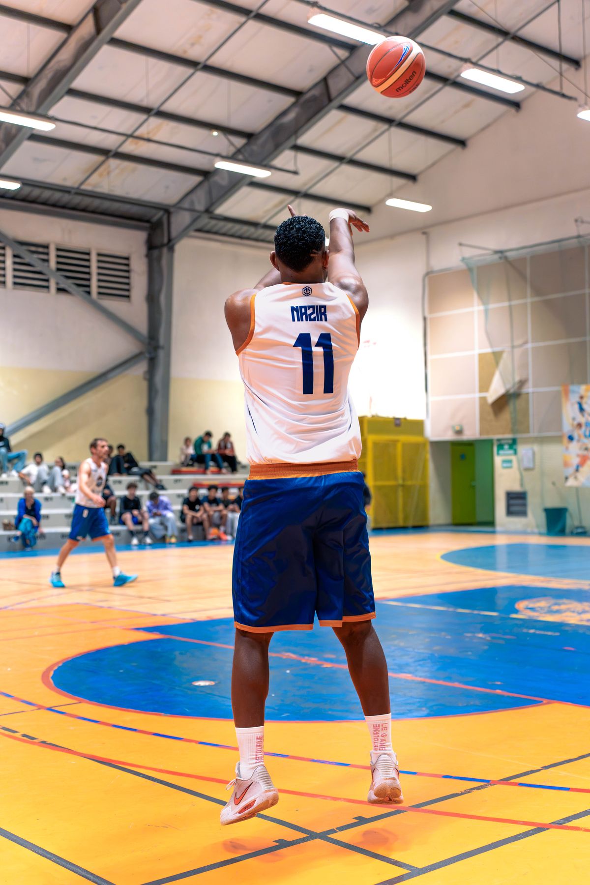 Kevin Nazir, numéro 11 du MBC, vu de dos en plein tir en suspension devant les tribunes ; le ballon s'élève sous la charpente du gymnase.