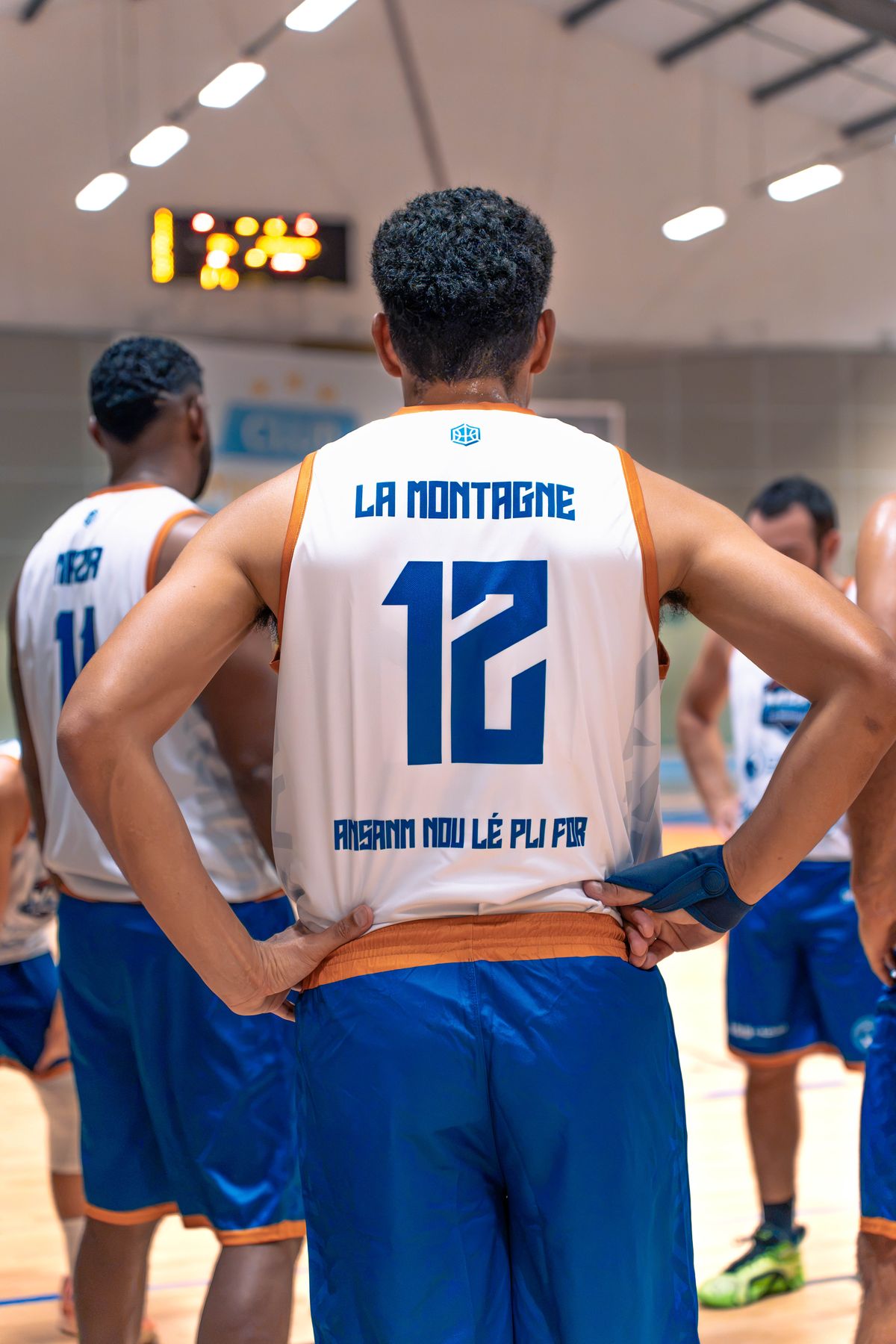 Le numéro 12 du MBC, de dos, mains sur les hanches : son maillot porte « La Montagne » et l'inscription créole « Ansanm nou lé pli for ». Au fond, le tableau d'affichage.