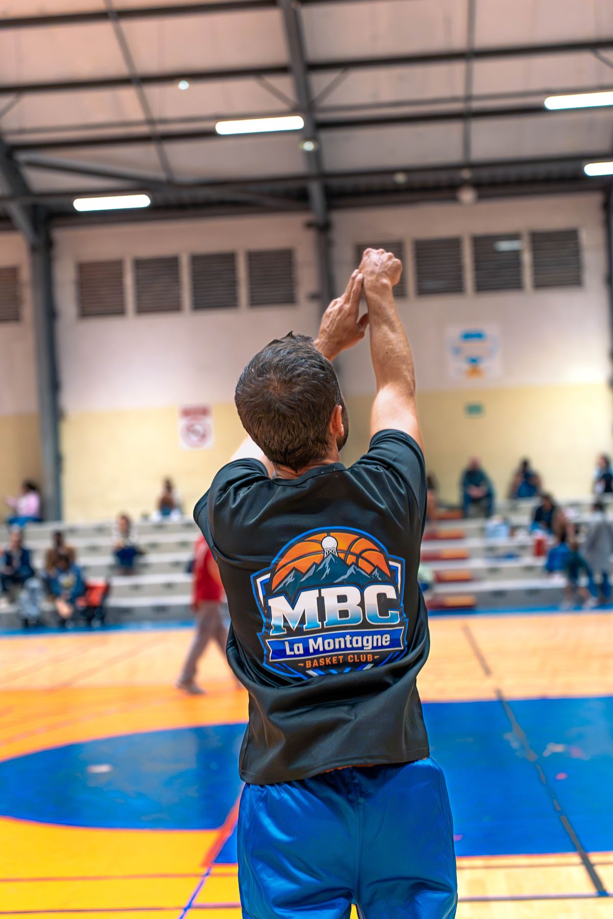 Un joueur du MBC en tee-shirt « MBC La Montagne Basket Club », vu de dos, bras tendus au bout de son tir, devant les tribunes du gymnase.
