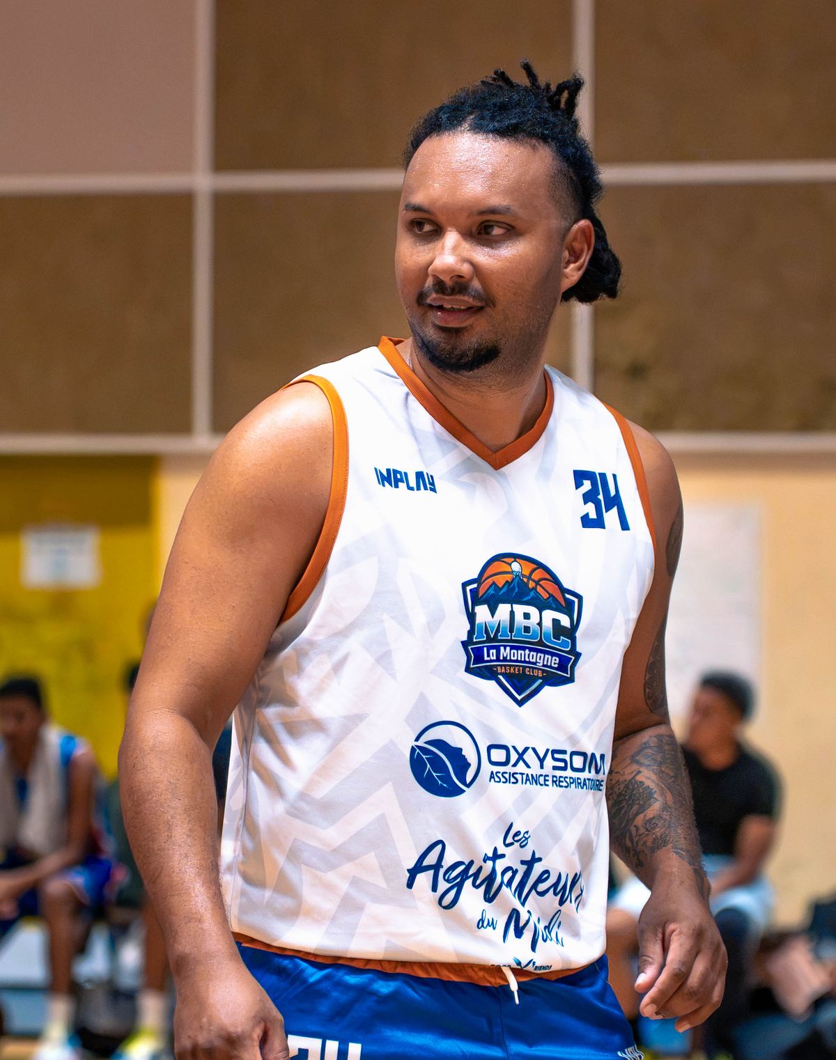 Maillot blanc 2026/2027 du MBC La Montagne, porté au gymnase de La Montagne à Saint-Denis (La Réunion) : la marque de l'équipementier InPlay en haut, le numéro 34, l'écusson du club au centre, le logo Oxysom sous l'écusson et la signature Les Agitateurs du Midi en bas de maillot.